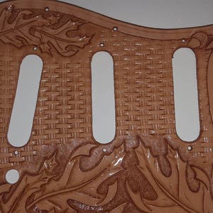 Puede incluir: Un pickguard de guitarra de cuero marr&oacute;n con un patr&oacute;n tejido y un dise&ntilde;o floral. El pickguard tiene tres recortes para pastillas.