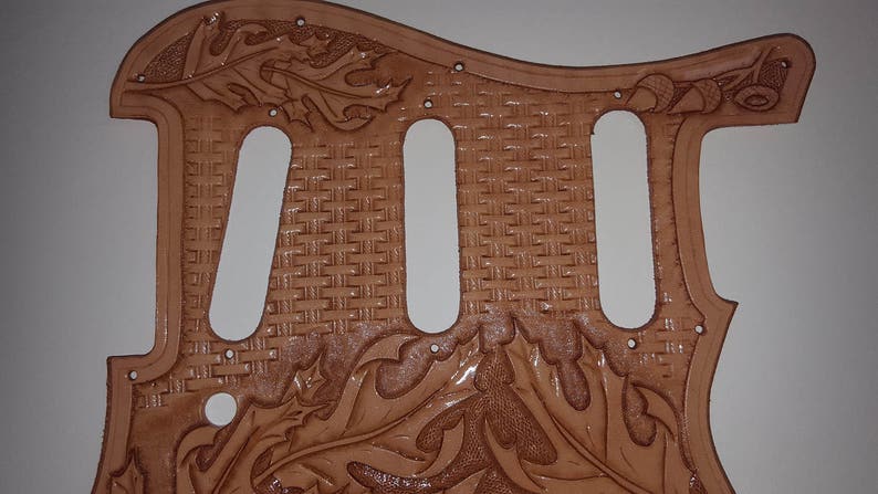 Puede incluir: Un pickguard de guitarra de cuero marr&oacute;n con un patr&oacute;n tejido y un dise&ntilde;o floral. El pickguard tiene tres recortes para pastillas.