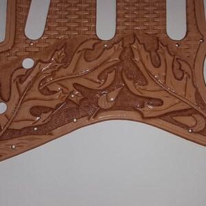 Puede incluir: Una placa de protecci&oacute;n de guitarra de cuero marr&oacute;n con un dise&ntilde;o floral tallado. La placa de protecci&oacute;n tiene un patr&oacute;n tejido en la secci&oacute;n superior.