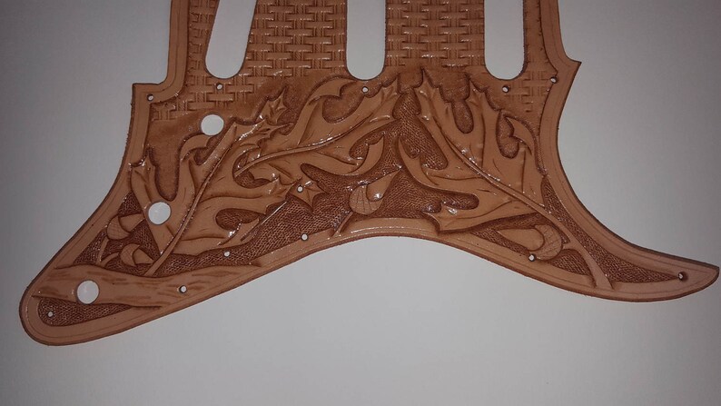 Puede incluir: Una placa de protecci&oacute;n de guitarra de cuero marr&oacute;n con un dise&ntilde;o floral tallado. La placa de protecci&oacute;n tiene un patr&oacute;n tejido en la secci&oacute;n superior.