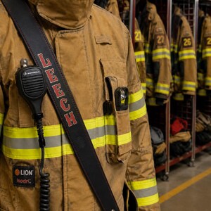 May include: A tan firefighter turnout coat with yellow and silver reflective stripes, featuring a black leather radio strap across the chest. The strap has the name LEECH embossed in red letters and holds a black Motorola radio microphone.