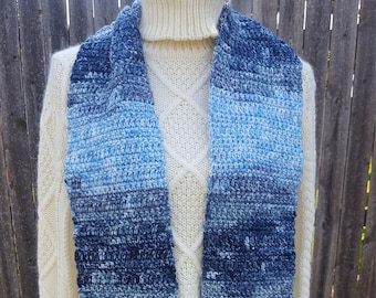 May include: A blue and white gradient crochet scarf with fringe. The scarf is draped over a cream-colored turtleneck sweater. The scarf's color transitions from light blue to dark blue.