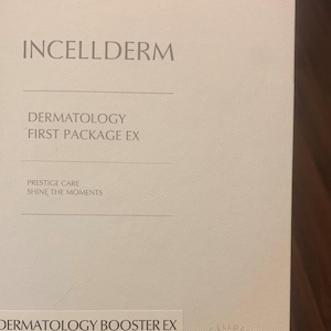 Op de afbeelding: Een witte doos met de tekst "INCELLDERM" in zwart bovenaan. De tekst "DERMATOLOGY FIRST PACKAGE EX" staat in zwart onder de bedrijfsnaam. De tekst "PRESTIGE CARE SHINE THE MOMENTS" staat in zwart onder de pakketnaam. De tekst "DERMATOLOGY BOOSTER EX" staat in zwart onderaan de doos.