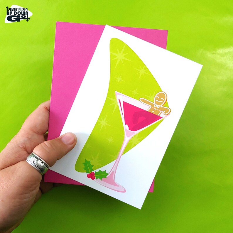 May include: A white card with a green and gold background featuring a pink martini glass with a gingerbread man inside. The martini glass has a holly sprig with red berries at the bottom. The card is folded in half and has a pink envelope.