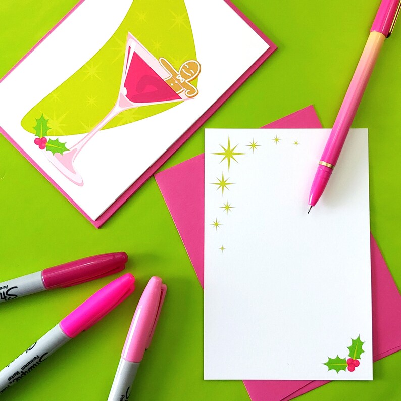 May include: A green and white Christmas card with a martini glass filled with red liquid and a gingerbread man inside. The card has a holly design and gold stars. A pink envelope and a pink pen are also in the image.