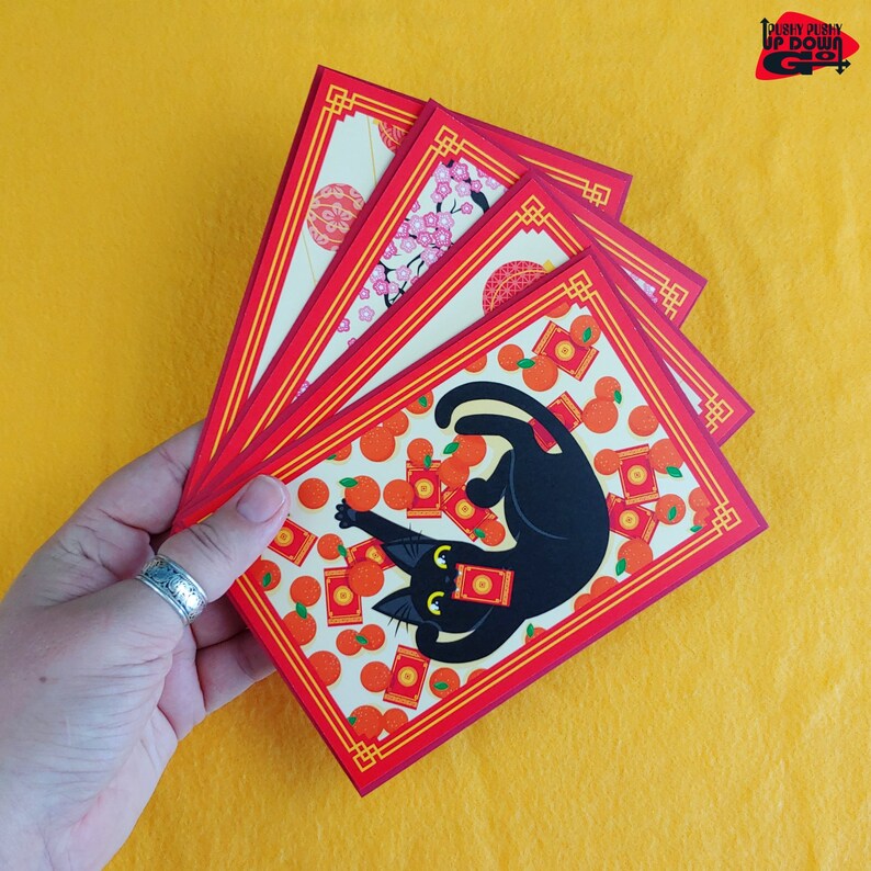 May include: A hand holds a fan of red and gold envelopes with a black cat illustration. The envelopes feature a repeating pattern of oranges and red packets, with a decorative border. The background is a solid yellow.