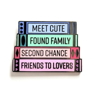 May include: A stack of four colorful enamel pins with the text "Meet Cute", "Found Family", "Second Chance", and "Friends to Lovers".