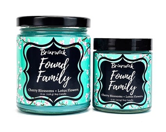 May include: Two soy candles in glass jars with black lids. The larger jar is 8 ounces and the smaller jar is 4 ounces. Both jars have a teal blue background with white cherry blossoms and the text "Briarwick Found Family Cherry Blossoms + Lotus Flowers" printed on the label.