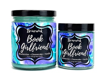 May include: Two soy candles in glass jars with black lids. The larger jar is 8 ounces and the smaller jar is 4 ounces. Both jars have a teal blue and purple marbled design with a black label that reads "Briarwick Book Girlfriend" and lists the scent as "Blueberries + Cheesecake + Vanilla".