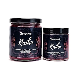 May include: Two glass candle jars with black lids. The larger jar is 8 ounces and the smaller jar is 4 ounces. Both jars are filled with a deep red candle wax and have a black label with white text that reads "Briarwick Raihu Desert Skies + Warmth + Saffron Officially Licensed".