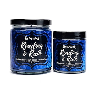 May include: Two blue soy candles with black lids and labels. The larger candle is 8 ounces and the smaller candle is 4 ounces. Both candles have the same label with the text "Briarwick Reading & Rain" and "Rain Water + Soft Linen + White Tea".