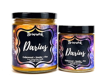 May include: Two soy candles in glass jars with black lids. The larger candle is 8 ounces and the smaller candle is 4 ounces. Both candles have a galaxy-themed label with the text "Briarwick Darius Cedarwood + Smoke + Fire Officially Licensed".
