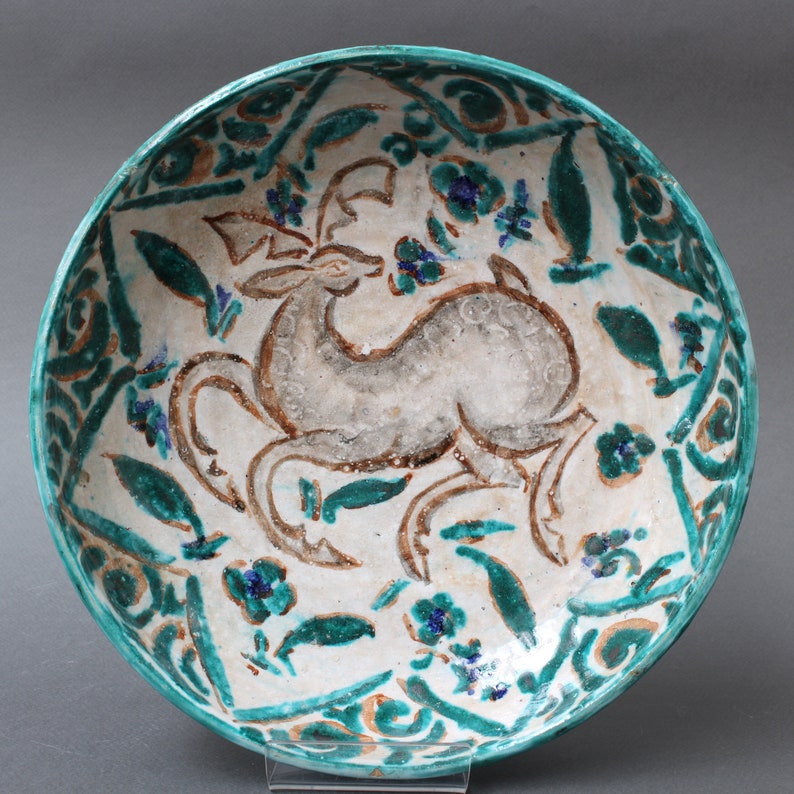 May include: A white ceramic bowl with a green and brown glaze. The bowl features a stylized deer design in the center, surrounded by floral motifs.