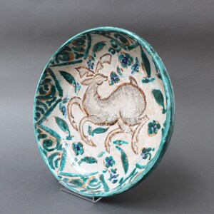 May include: A white ceramic bowl with a green and blue floral design and a stylized deer in the center. The bowl is on a clear stand.