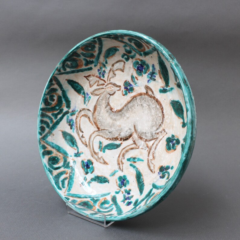 May include: A white ceramic bowl with a green and blue floral design and a stylized deer in the center. The bowl is on a clear stand.