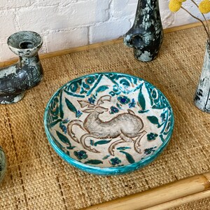 May include: A turquoise ceramic bowl with a white and gray floral design and a stylized animal in the center. The bowl is on a woven bamboo surface.