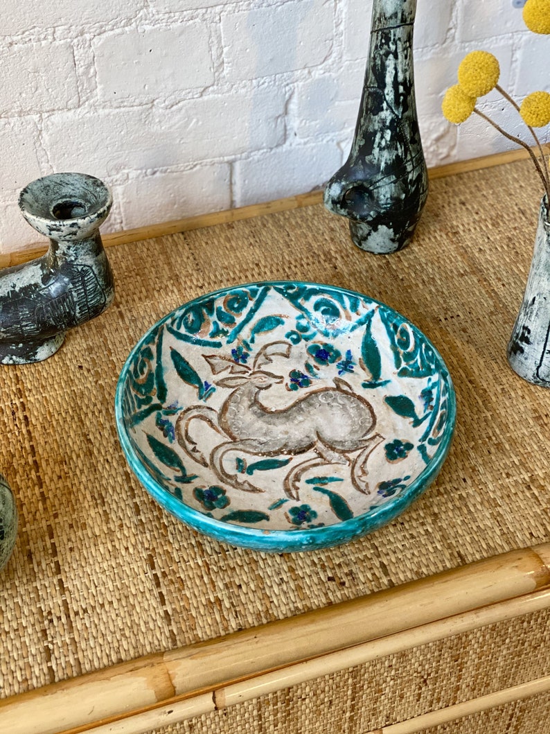 May include: A turquoise ceramic bowl with a white and gray floral design and a stylized animal in the center. The bowl is on a woven bamboo surface.