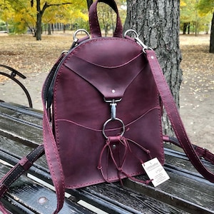 Puede incluir: Una mochila de cuero burdeos de forma redonda con un detalle de anillo plateado. La mochila tiene un asa superior y correas ajustables. La bolsa tiene una pequeña etiqueta adjunta que dice "Cuir hecho a mano".