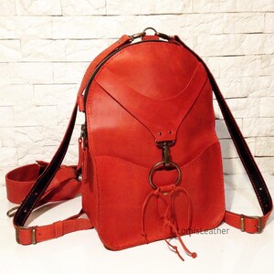 Puede incluir: Una mochila de cuero rojo brillante con un dise&ntilde;o &uacute;nico que presenta un gran anillo de metal y un nudo decorativo. La mochila tiene un cierre de cremallera y correas ajustables.