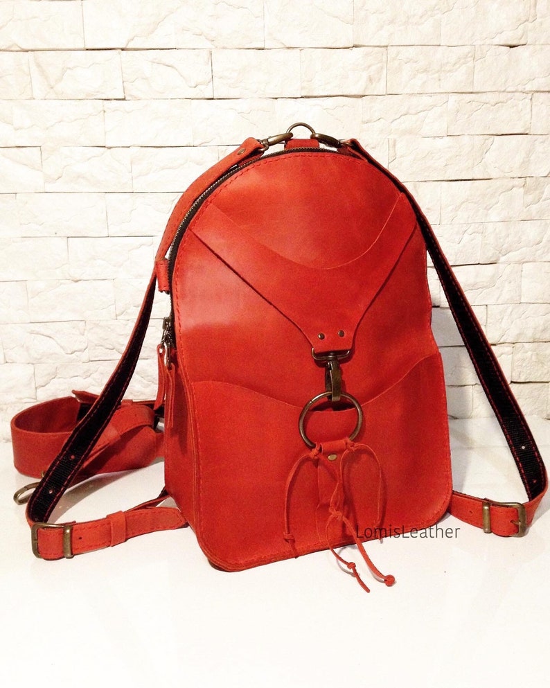 Puede incluir: Una mochila de cuero rojo brillante con un dise&ntilde;o &uacute;nico que presenta un gran anillo de metal y un nudo decorativo. La mochila tiene un cierre de cremallera y correas ajustables.