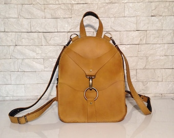May include: A mustard yellow leather backpack with a large metal ring and buckle closure. The backpack has adjustable straps and a small pocket on the front.