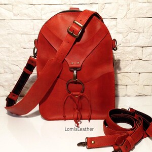 May include: A red leather crossbody bag with a unique design featuring a large metal ring and a knotted leather strap. The bag has a zipper closure and a long adjustable strap.