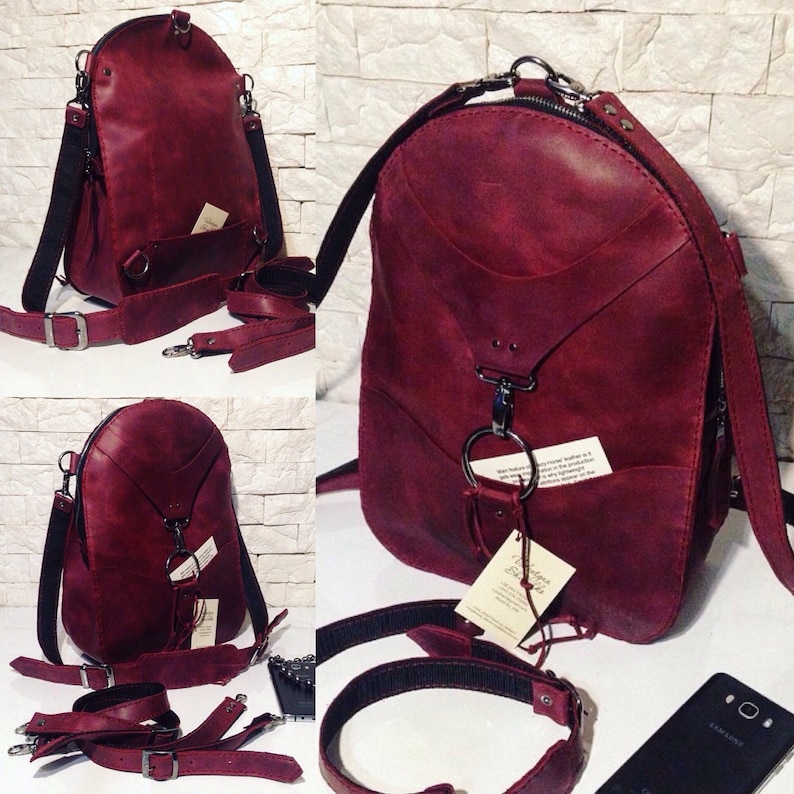 Puede incluir: Una mochila de cuero burdeos con una sola correa y una hebilla plateada. La mochila tiene cierre de cremallera y un peque&ntilde;o bolsillo en la parte delantera. La mochila se muestra sobre un fondo de pared de ladrillo blanco.