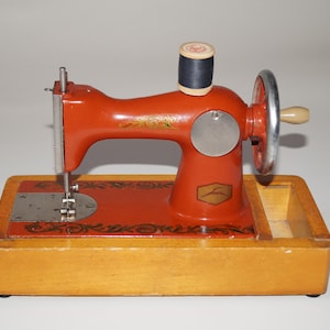 Peut inclure: Une machine à coudre rouge vintage avec une base en bois et une bobine de fil noire. La machine a un volant argenté et un petit compartiment de rangement. Des motifs floraux décoratifs sont visibles.