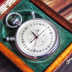 May include: A classic stopwatch with a silver casing and a white face, presented in a wooden box with green velvet lining. The stopwatch has black hands and a red second hand, with numbers around the dial. A small metal tool is next to the box.