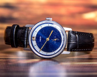 Puede incluir: Un reloj de pulsera vintage con esfera azul oscuro y agujas doradas. El reloj tiene una caja plateada y una correa de cuero negro. La palabra "POBEDA" está impresa en la esfera. Fondo borroso.