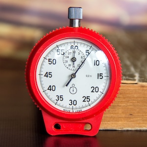 Op de afbeelding: Een rode stopwatch met een witte wijzerplaat en zwarte cijfers. De stopwatch heeft een kleine wijzerplaat aan de bovenkant en een grotere met markeringen voor seconden. De tekst "MADE IN USSR" is zichtbaar.