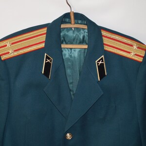 May include: A teal military jacket with gold and red shoulder boards featuring stars. The jacket has black collar tabs with gold accents and a gold button. The jacket is hanging on a wooden hanger.