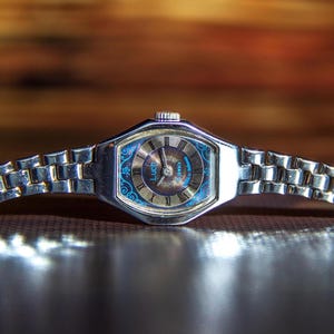May include: A silver-toned wristwatch with a metallic link bracelet. The watch face features a dark brown center, a blue border with intricate floral patterns, and the text Luch. It has silver hands and Roman numeral hour markers.
