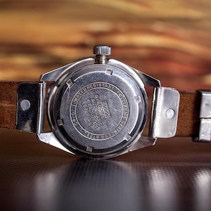Puede incluir: Primer plano de la parte trasera de un reloj de pulsera vintage con una correa de cuero marr&oacute;n. La caja del reloj es plateada con texto grabado, incluyendo "Water Resistant" y "Stainless Steel". La correa est&aacute; sujeta con piezas met&aacute;licas plateadas.