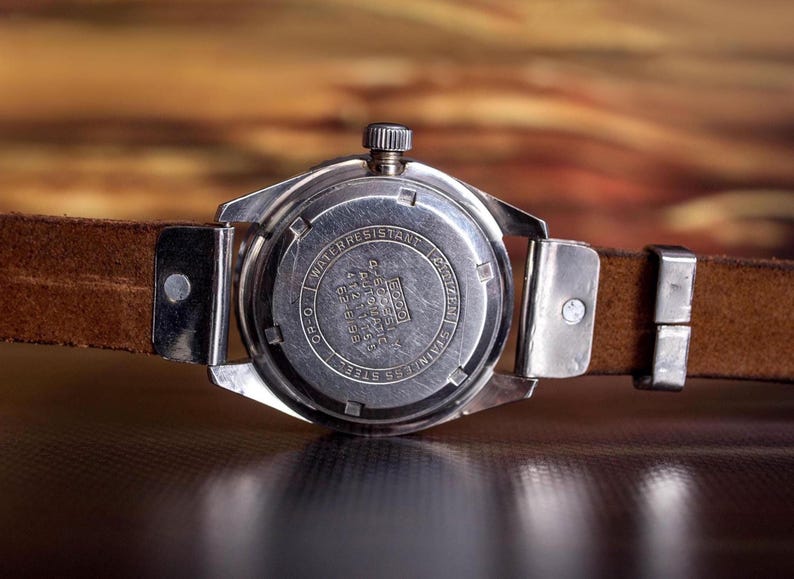 Puede incluir: Primer plano de la parte trasera de un reloj de pulsera vintage con una correa de cuero marr&oacute;n. La caja del reloj es plateada con texto grabado, incluyendo "Water Resistant" y "Stainless Steel". La correa est&aacute; sujeta con piezas met&aacute;licas plateadas.