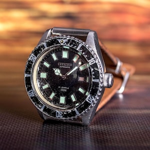 Puede incluir: Un reloj de pulsera autom&aacute;tico Citizen vintage con esfera negra y marcadores luminosos. El reloj tiene una caja de acero inoxidable, un bisel giratorio y una correa de cuero marr&oacute;n. La esfera del reloj muestra el texto "Citizen Automatic" y "150M".