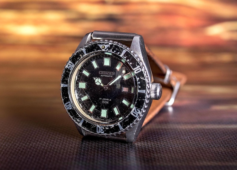 Puede incluir: Un reloj de pulsera autom&aacute;tico Citizen vintage con esfera negra y marcadores luminosos. El reloj tiene una caja de acero inoxidable, un bisel giratorio y una correa de cuero marr&oacute;n. La esfera del reloj muestra el texto "Citizen Automatic" y "150M".