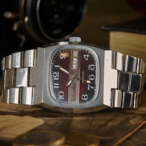 Op de afbeelding: Een vintage zilveren polshorloge met een rechthoekige wijzerplaat en een bordeauxrode wijzerplaat. Het horloge heeft een zilveren band en geeft de dag en datum weer. Het horloge staat tegen een achtergrond van een camera en een boek.