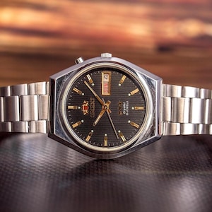 May include: A silver-tone analog wristwatch with a textured black dial, gold-tone hands, and hour markers. The watch features a day-date window at the three o'clock position, the Orient logo, and a stainless steel link bracelet with a fold-over clasp.