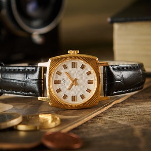 May include: A vintage gold-toned wristwatch with a square face and a black leather band. The watch face is white with gold hands and hour markers. The watch is displayed on a wooden surface with coins and a book in the background.