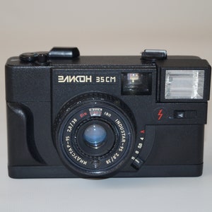 May include: Black vintage camera with the text "ЗЕЛКОН 35CM" in gold lettering. The camera has a lens, flash, and various dials and settings. The lens is labeled "INDUSTAR" and has measurement markings.