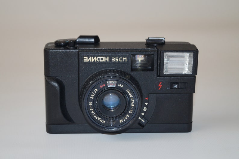 May include: Black vintage camera with the text "ЗЕЛКОН 35CM" in gold lettering. The camera has a lens, flash, and various dials and settings. The lens is labeled "INDUSTAR" and has measurement markings.