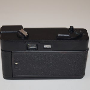 May include: A black vintage camera with a textured body. The camera has a flash mount, a lens, and various dials and buttons on top. The back of the camera has a textured surface. The camera is set against a white background.