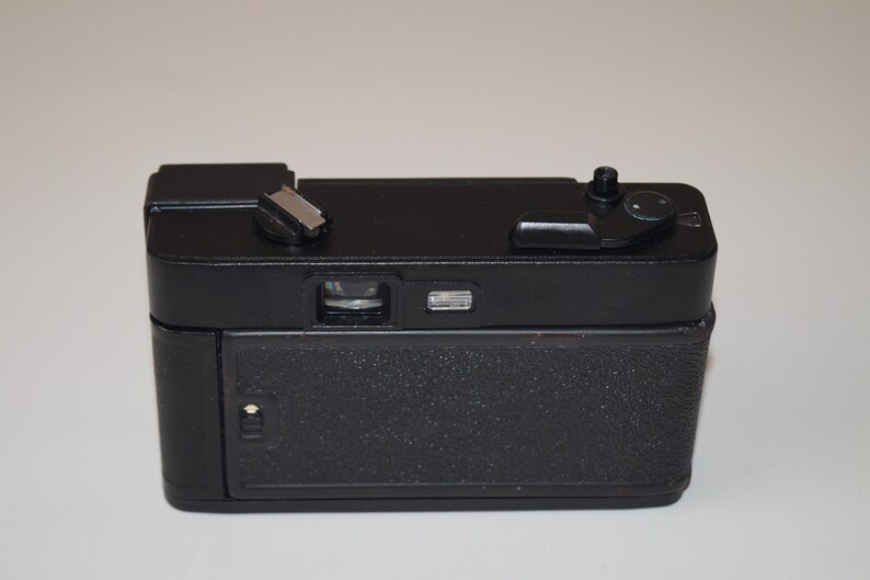 May include: A black vintage camera with a textured body. The camera has a flash mount, a lens, and various dials and buttons on top. The back of the camera has a textured surface. The camera is set against a white background.