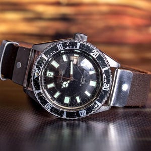 Puede incluir: Un reloj de estilo vintage con esfera negra y bisel giratorio. El reloj tiene marcadores y agujas luminosas, caja de acero inoxidable y correa de cuero marr&oacute;n. El fondo est&aacute; borroso.