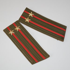 May include: Pair of olive green military shoulder boards with two gold star emblems and red stripes. The epaulettes are rectangular and feature a woven texture. The background is plain white.