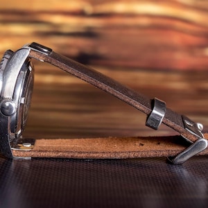Puede incluir: Un reloj de pulsera de acero inoxidable con una correa de cuero marr&oacute;n. La esfera del reloj es negra con detalles plateados. La correa de cuero es de color marr&oacute;n medio y tiene un cierre de hebilla.