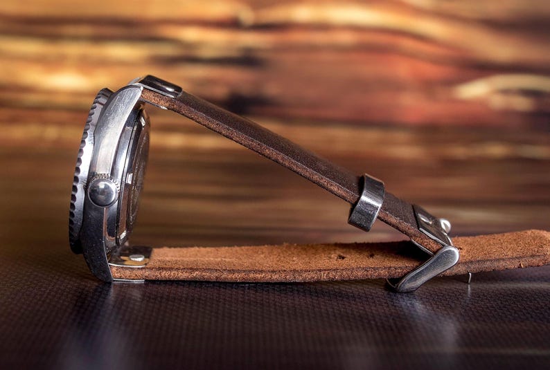 Puede incluir: Un reloj de pulsera de acero inoxidable con una correa de cuero marr&oacute;n. La esfera del reloj es negra con detalles plateados. La correa de cuero es de color marr&oacute;n medio y tiene un cierre de hebilla.