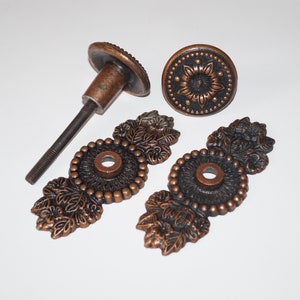 Può includere: Quattro pezzi di ferramenta per mobili antichi in bronzo. Include due maniglie ornate con accenti floreali, un pomello rotondo con un motivo floreale e un pomello con un'asta filettata. L'hardware è di un caldo colore rame.