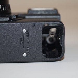May include: Close-up of a black camera body with a lens and open battery compartment. The camera has a textured surface and the text "MADE IN USSR" is visible. The camera is on a white surface.